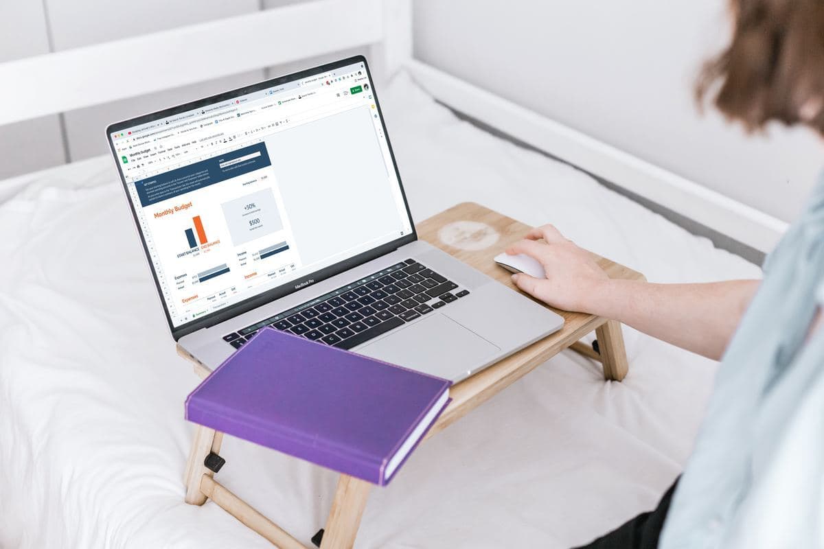 A woman sitting on a bed, using a laptop with a notebook open beside her.